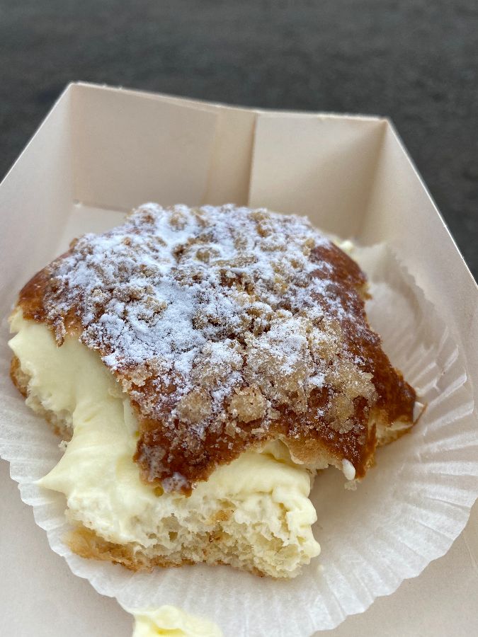 종이 트레이에 담긴 페이스트리의 클로즈업 사진. 슈가파우더가 뿌려진 바삭한 겉면과 부드러운 크림 속이 선명하게 보여 식욕을 자극합니다.