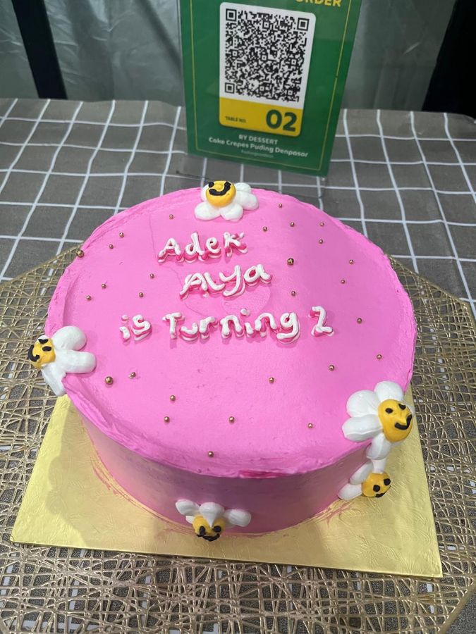 밝은 핑크색 생일 케이크 위에 'Adek Aya is Turning 2'라는 문구와 노란색 꽃 장식이 있는 모습